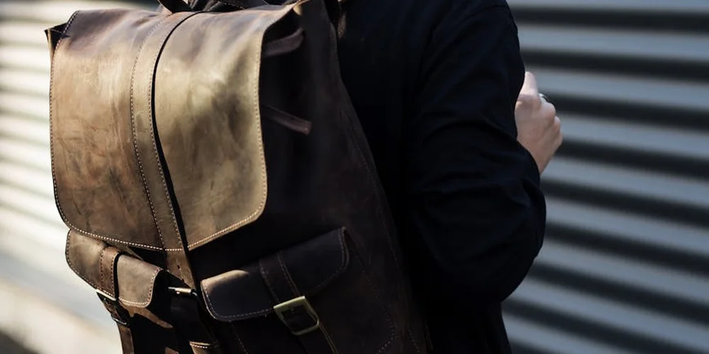 Brown leather backpack worn by a person in a black jacket, standing in front of a corrugated metal wall.